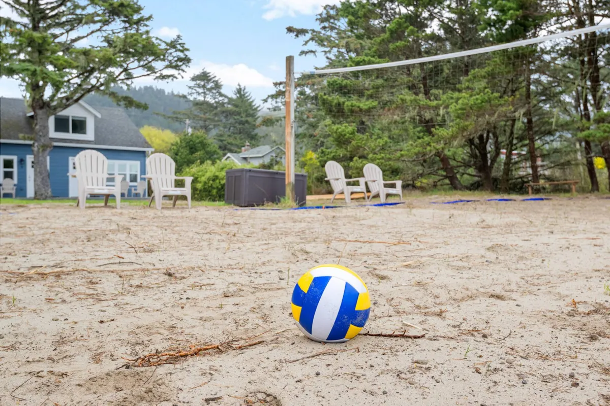 An inviting outdoor space centered around a volleyball court, with a net strung across sandy ground, surrounded by beach chairs and tall trees. The setup radiates a playful, sociable energy and hints at both relaxation and friendly competition.