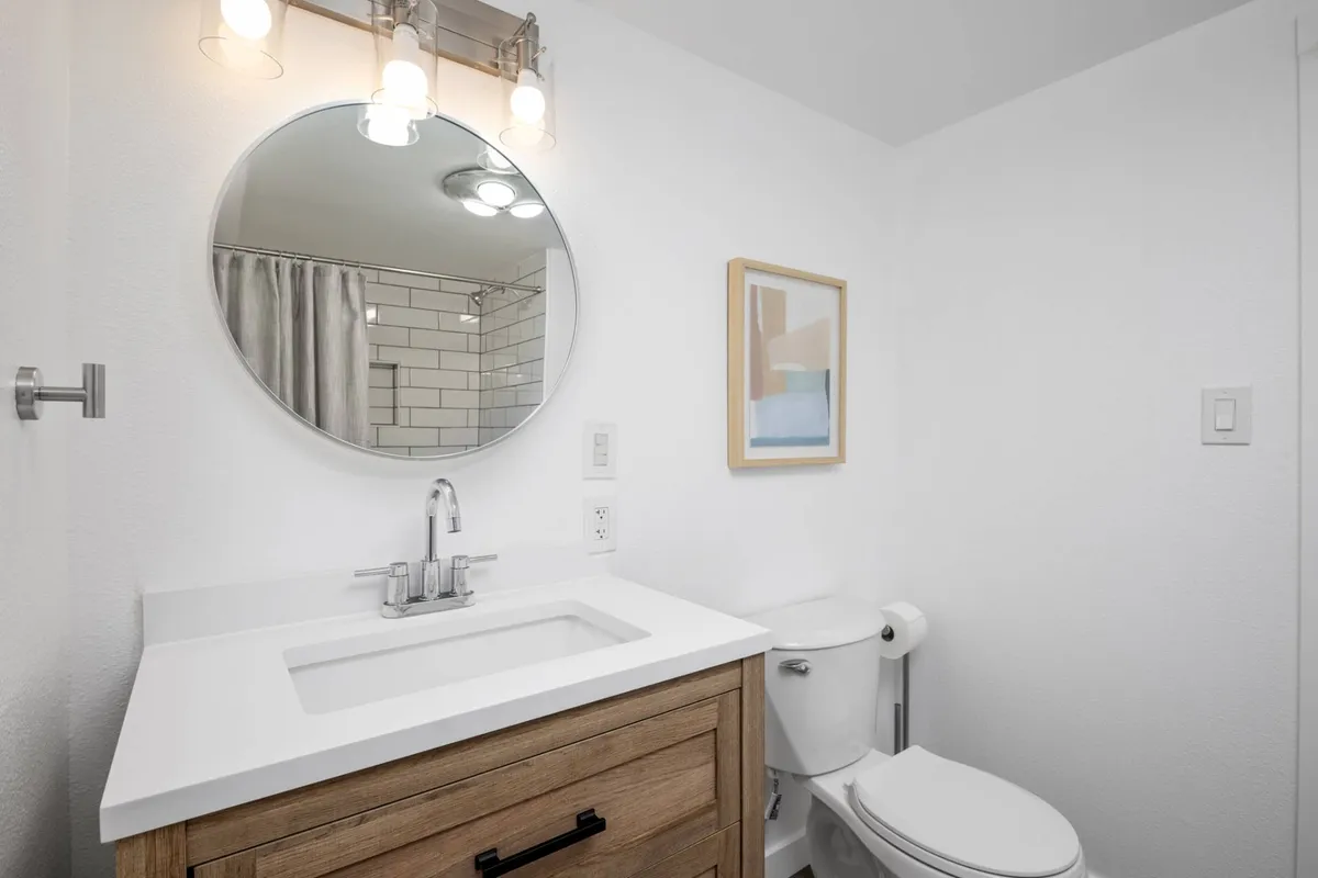 A stylish bathroom interior featuring a round mirror above a wooden vanity, accentuated by framed artwork. The space feels fresh and intimate, ideal for morning routines and self-care.