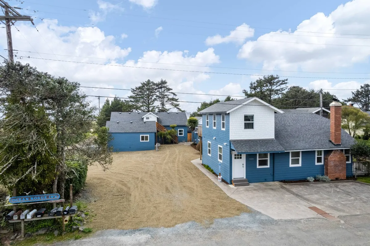 A broad exterior view showing multiple blue houses surrounded by trees and a gravel driveway. The open yard space and natural surroundings hint at group activities, outdoor games, and easy access to explore the area.
