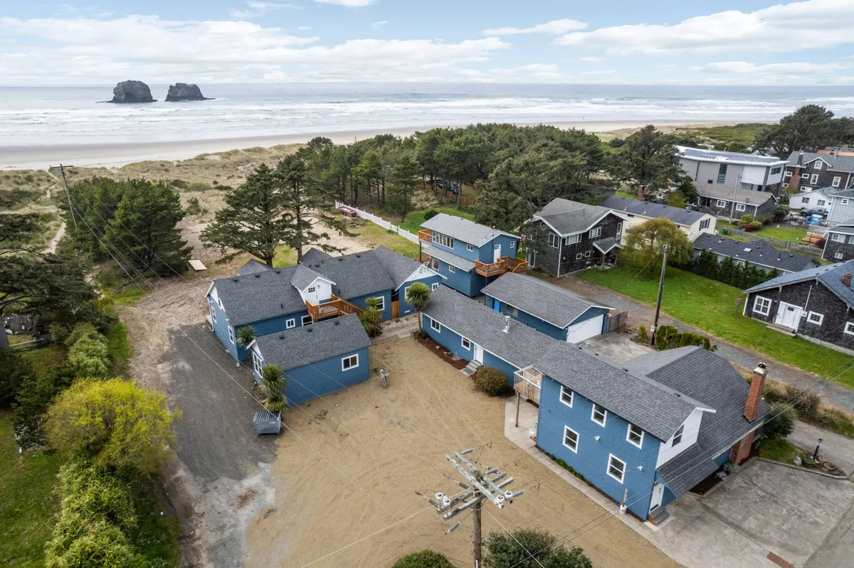 This image captures a serene stretch of beach with Twin Rocks visible offshore, a sandy beach area ideal for family activities, and a welcoming cluster of blue houses nestled by green trees. The scene showcases the proximity of the property to the ocean, the iconic coastal rock formations, and the immediate access to sand and surf.