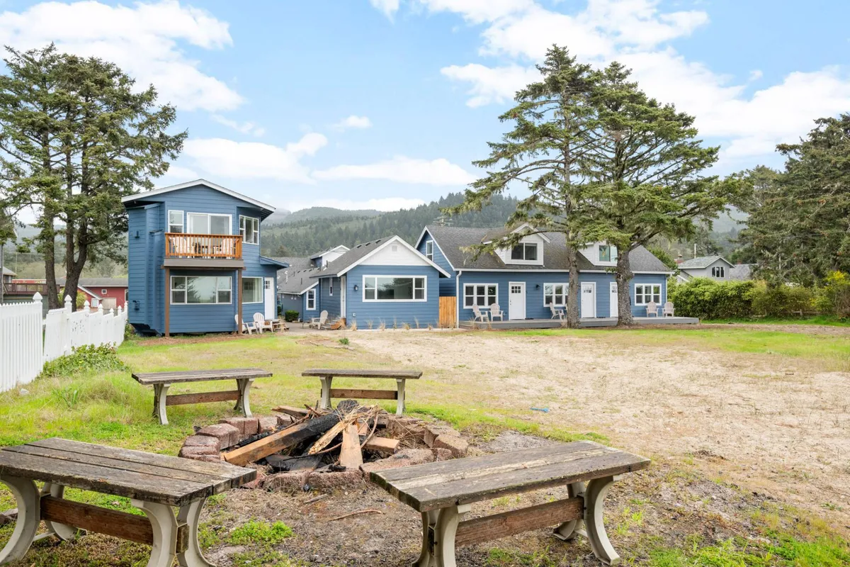 An exterior scene with a cozy fire-pit surrounded by blue houses and trees, inviting guests to gather for outdoor storytelling or gatherings. The setup encourages community and warmth, especially during sunset or cool evenings.