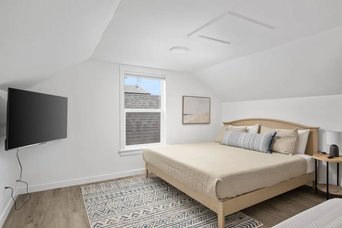 A spacious bedroom with a large bed dressed in neutral bedding, a wall-mounted TV, tasteful artwork, and a window streaming in natural light. The environment is both restful and welcoming, perfect for unwinding after a lively day.