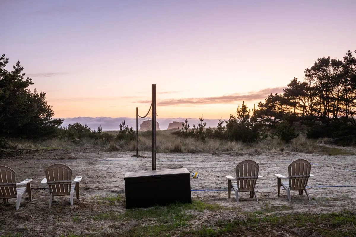 A broad exterior shot that includes Adirondack chairs, a volleyball net set up on sand, an ocean view, and a glowing sunset sky. The scene is both serene and lively, offering visual cues for relaxation, play, and enjoying the coastal setting.