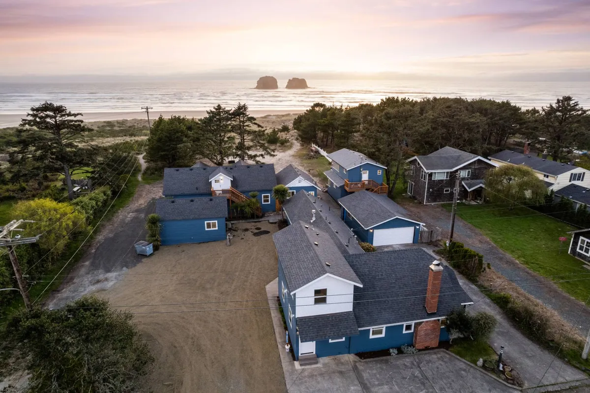 This image captures a scenic stretch of beach with the ocean, rock formations, and adjacent houses framed by trees. The sandy area is expansive and inviting, suggesting plenty of space for outdoor activities or pets to play, and the houses' proximity emphasizes direct beach access.