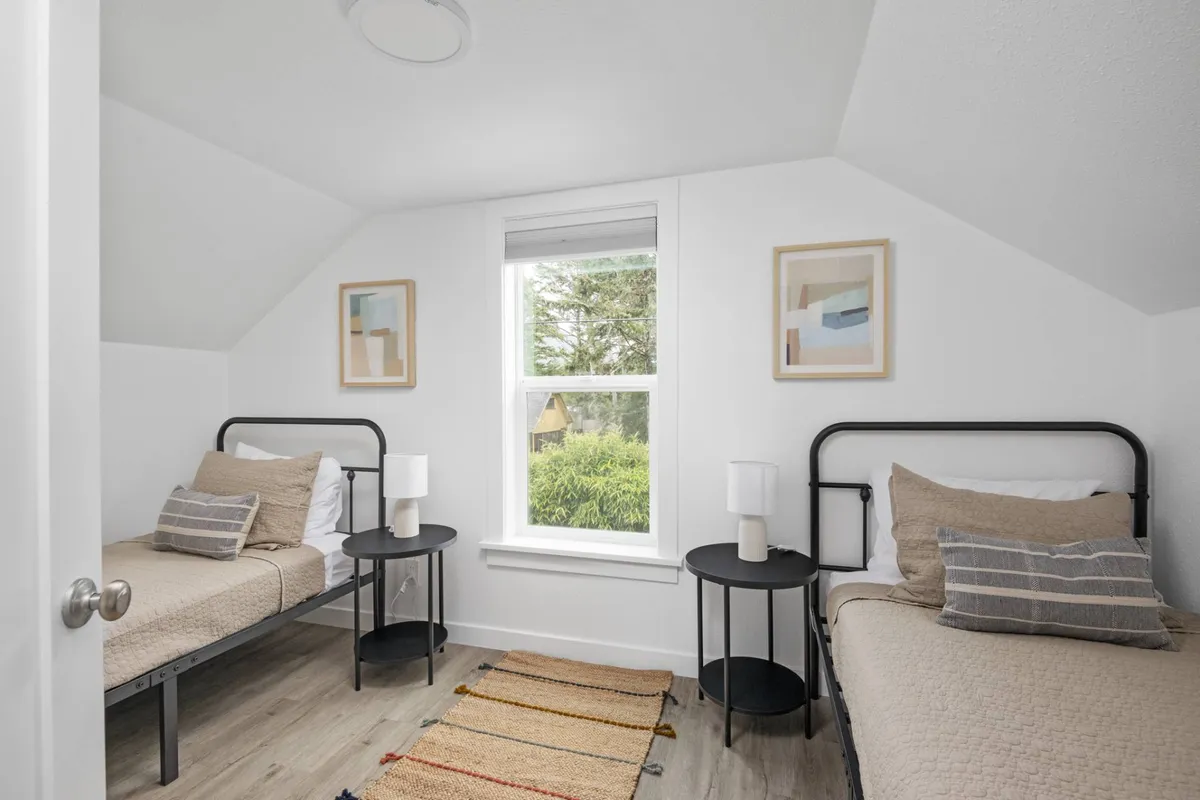 A serene bedroom featuring two twin beds, a window with natural light, decorative artwork, and cozy nightstands with lamps. The room feels inviting for children or teens, and encourages comfortable relaxation.