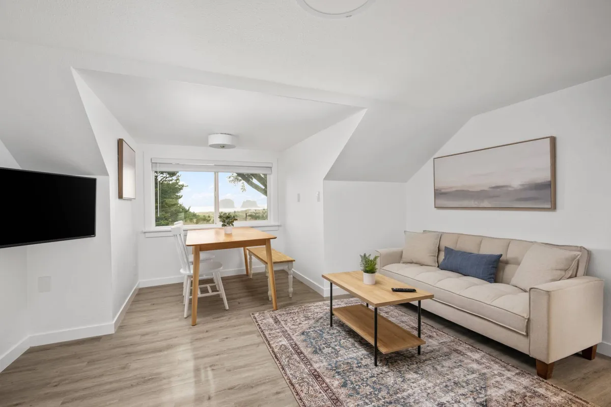 The image showcases a bright, inviting living space with a plush sofa, a charming dining table set for meals, a cozy rug, and large windows that frame an ocean view. The room feels open and airy, perfect for relaxing, dining, or gathering with friends and family, with natural light pouring in and a hint of the stunning Oregon coast just beyond the glass.