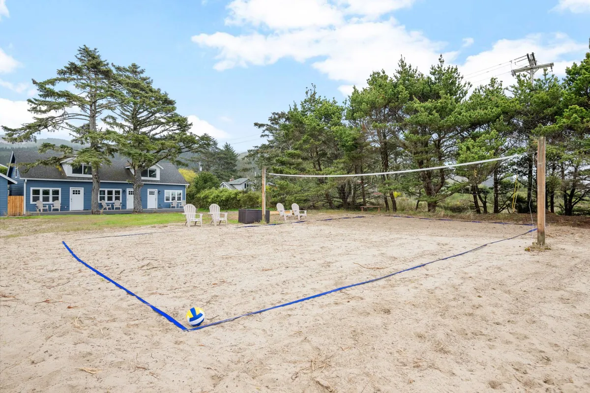 A playful exterior scene featuring a sand volleyball court bordered by Adirondack chairs and tall trees, inviting guests to gather for games, relaxation, and socializing. The area is clearly designed for family play and community bonding, echoing the lively atmosphere described in the narrative.
